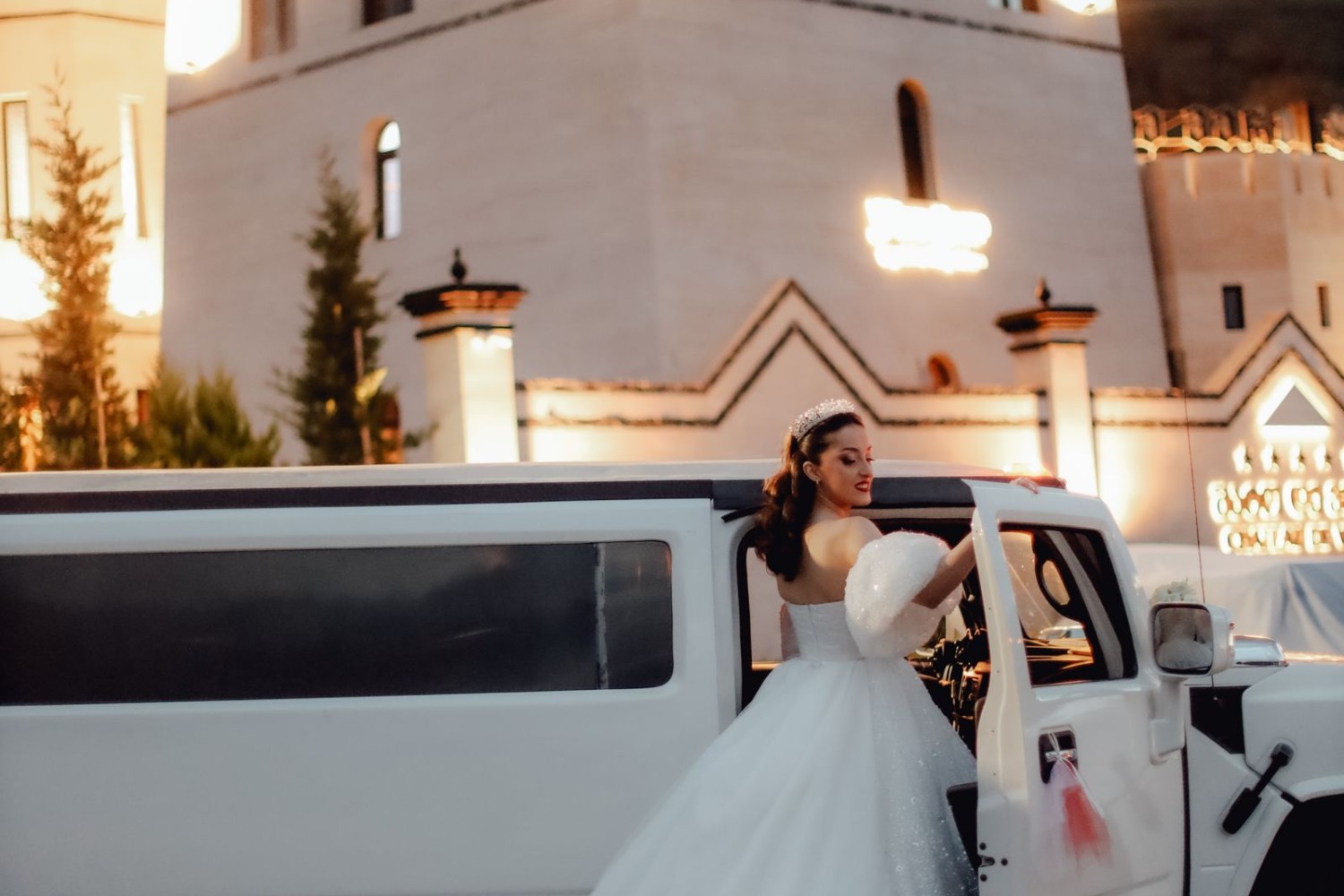 Bride beside a limousine outside an illuminated venue at twilight